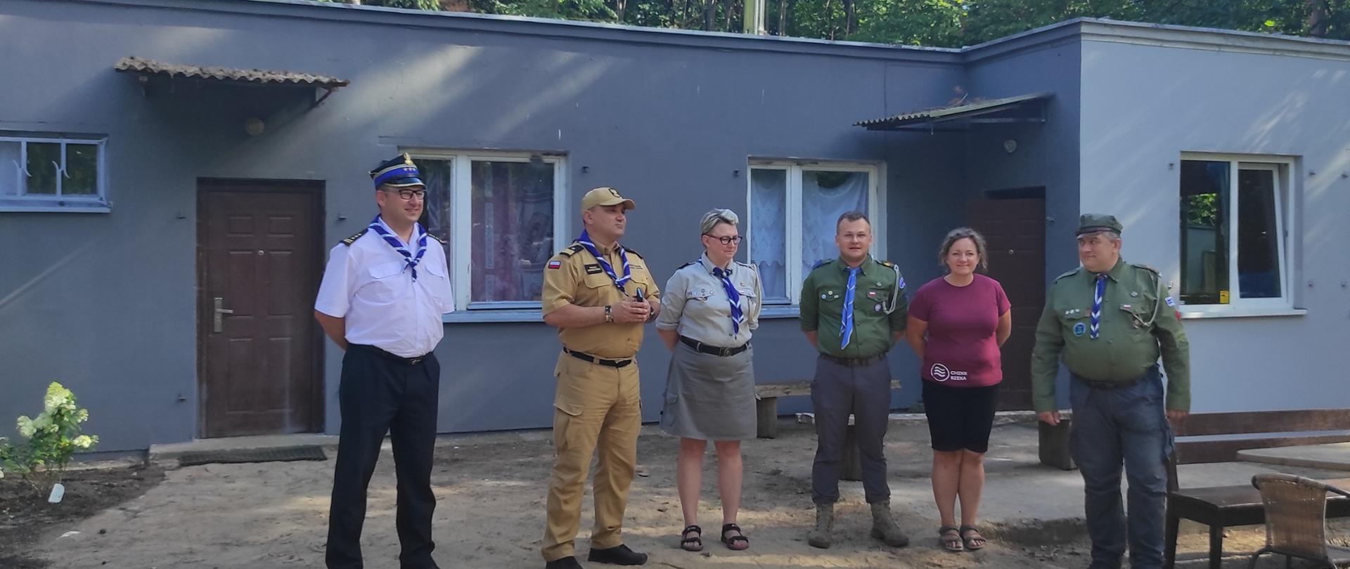 Przed szarym budynkiem stoją komendanci wojewódzcy straży pożarnej i czterech przedstawicieli harcerstwa. W oddali zielone drzewa.