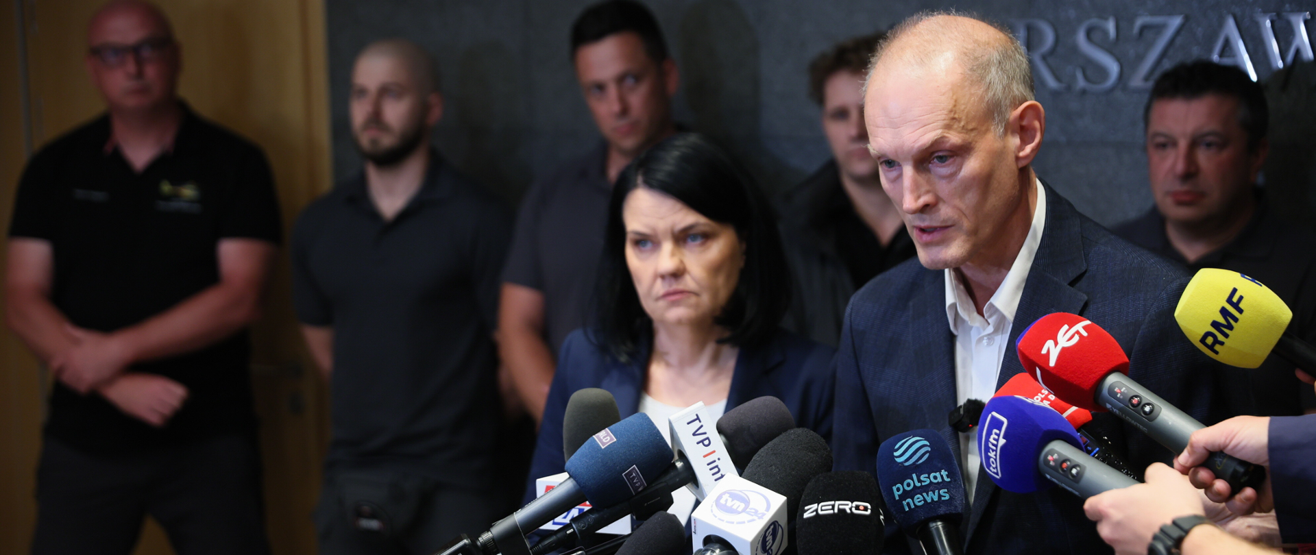 Wiceminister Zajączkowski przemawia podczas konferencji prasowej stoi przed licznymi mikrofonami z logo polskich stacji telewizyjnych i radiowych. Za nim stoi wiceminister zdrowia Katarzyna Kacperczyk oraz kilku mężczyzn. W tle widoczna jest ściana z napisem „Warszawa”.