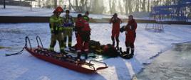 Strażacy przygotowujący sprzęt nad brzegiem zalewu do ćwiczeń z ratownictwa lodowego. Ubrani w ubrania do pracy w wodzie przygotowują do użycia deskę lodową
