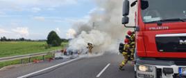 Pożar auta na S5. Jezdnia trasy S5 w kierunku Poznania. Na pasie awaryjnym stoi palący się samochód osobowy. Wokół niego silne zadymienie. Z prawej strony w kłębach dymu strażak gaszący pożar. Widoczne są płomienie. Na pierwszym planie wóz straży pożarnej i dwóch strażaków, którzy wyjmują ze skrytek sprzęt potrzebny do działań.
