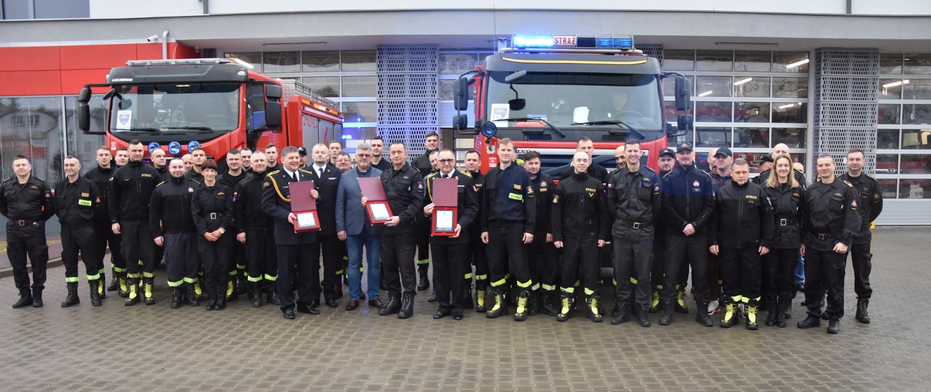 Wszyscy funkcjonariusze z emerytami na tle samochodów pożarniczych przed wrotami JRG