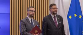Na tle ściany z cienkich pionowych listewek stoją wiceminister Szeptycki i minister Kulasek trzymający w rękach czerwoną teczkę, za nimi flagi Polski i UE.