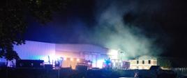 zdjęcie wykonane jest porą nocną, na środku zdjęcia widnieją
oświetlone hale
widać dwa samochody ratowniczo-gaśnicze z ustawionymi reflektorami
