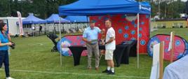 Piłkarz odbiera nagrodę od mężczyzny w koszuli wizytowej z krótkim rękawem. Za nimi namiot wystawowy w kolorach niebiesko - czerwonych. W tle inne namioty, drzewa.
