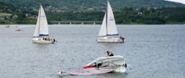 Widok z przodu na taflę jeziora. Na pierwszym planie przewrócona żaglówka. W tle dwie kolejne, drogowa estakada i góry. 
