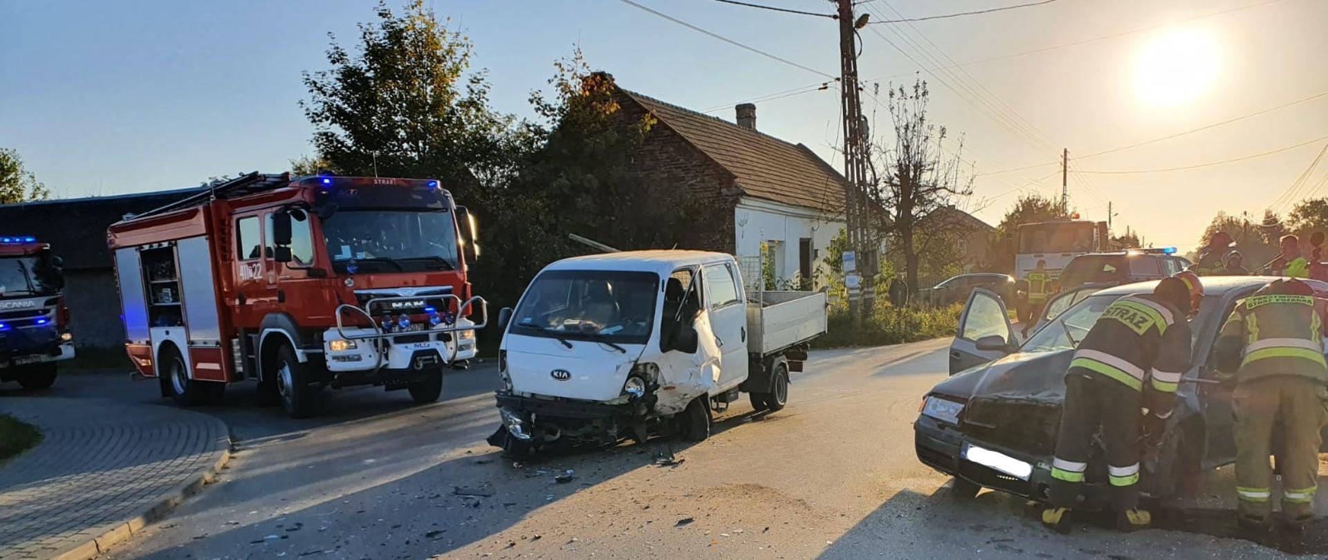 Zdjęcie przedstawia dwa uszkodzone pojazdy blokujące drogę powiatową. Na zdjęciu widać strażaków, którzy pracują przy odłączaniu akumulatora w samochodzie osobowym. Po lewej stronie stoją zaparkowany pojazdy ratowniczo-gaśnicze z włączonymi błyskowymi światłami alarmowymi w kolorze niebieskim.