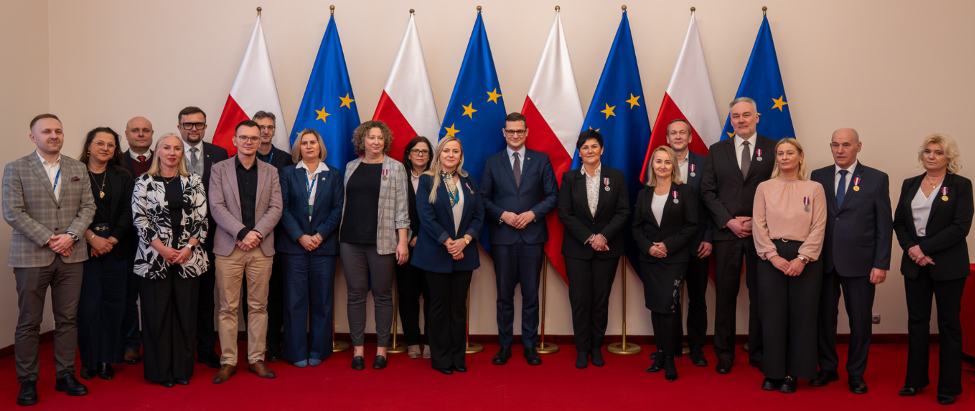 Zdjęcie grupowe uczestników uroczystości w Kujawsko-Pomorskim Urzędzie Wojewódzkim. Na środku stoi Wojewoda Michał Sztybel w otoczeniu grupy osób odznaczonych (kobiet i mężczyzn), którzy prezentują przypięte medale. Wszyscy stoją na tle flag Polski i Unii Europejskiej oraz godła państwowego.