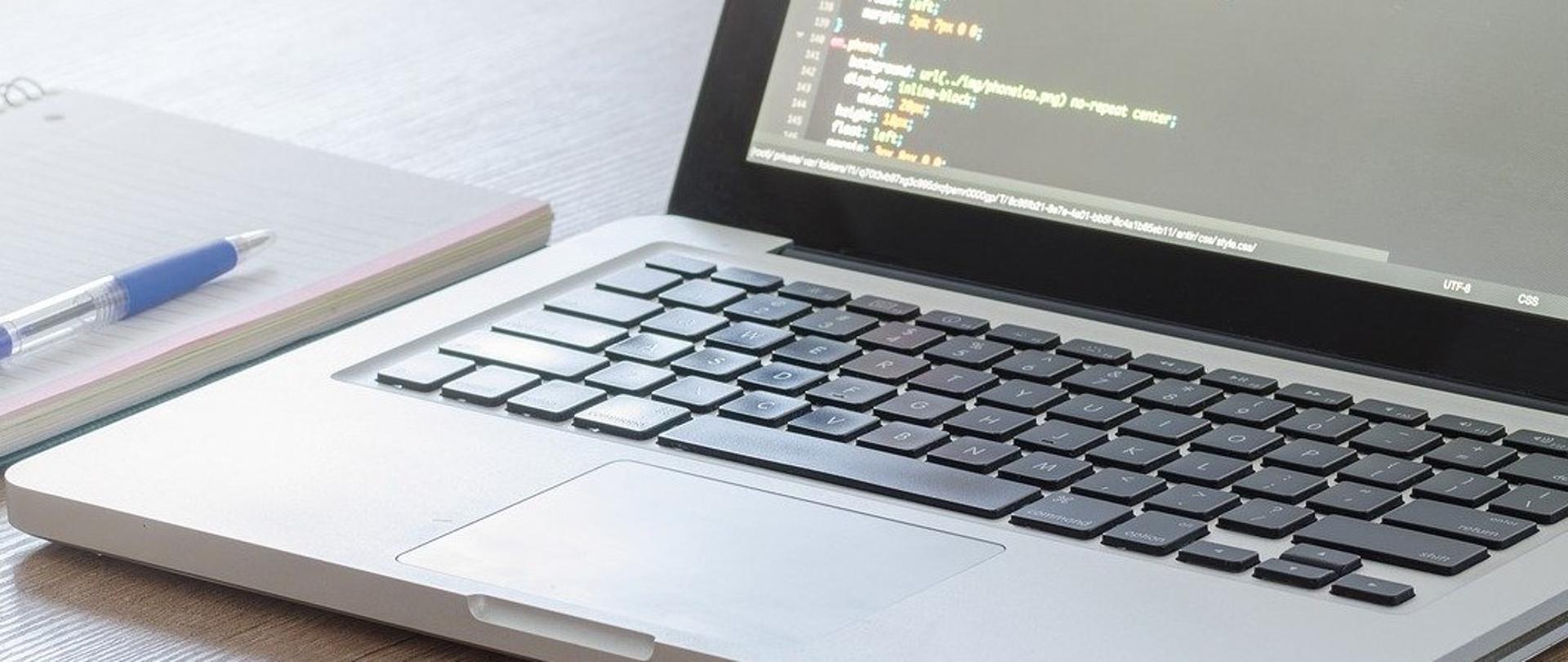 Laptop z otwartym edytorem kodu, obok notes i długopis na biurku.