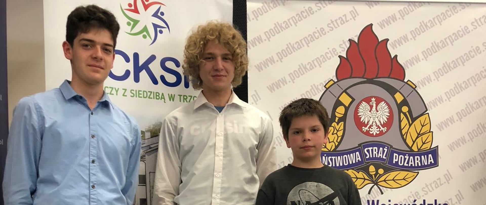 Na zdjęciu widać trzech uczestników eliminacji Ogólnopolskiego Turnieju Wiedzy Pożarniczej. Z tyłu za nimi loga różnych firm oraz Państwowej Straży Pożarnej. 