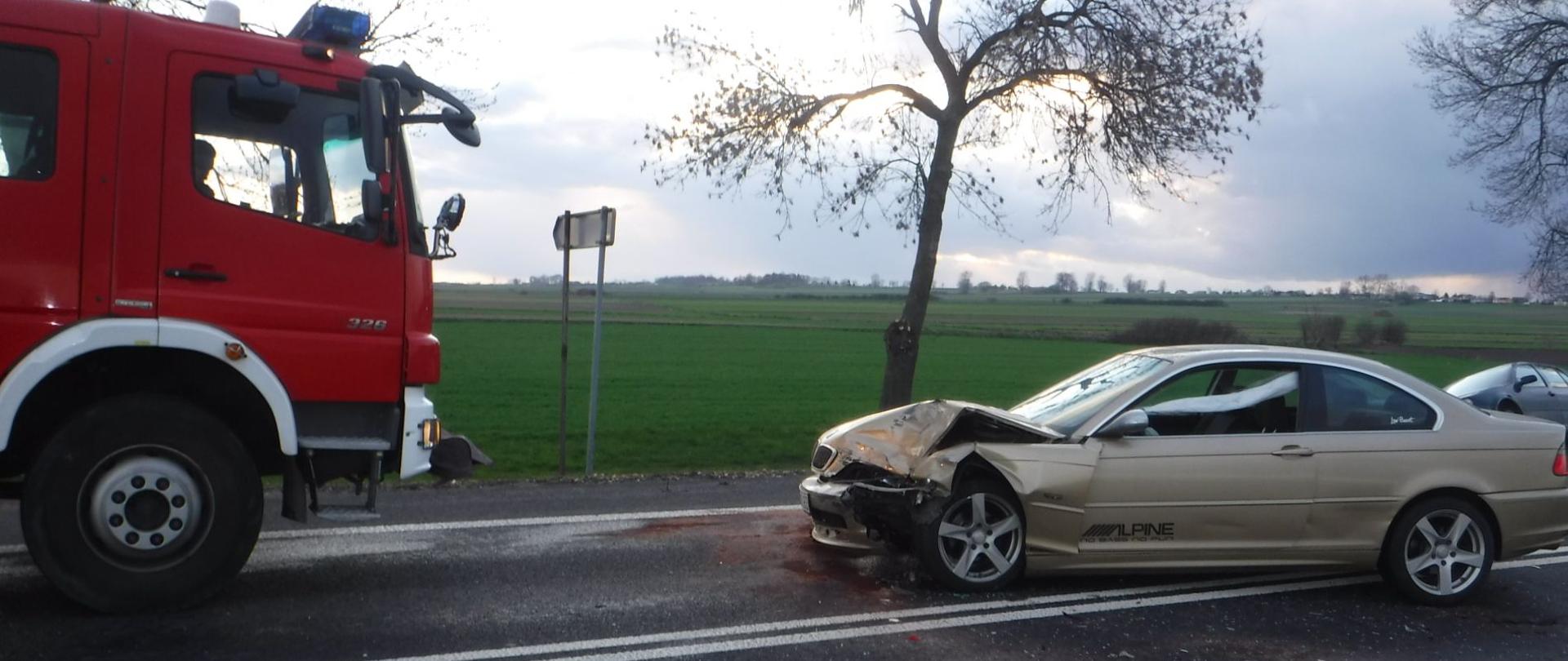 Na zdjęciu widać rozbity złoty samochód osobowy stojący na drodze oraz drugi rozbity ciemny samochód osobowy znajdujący się częściowo w rowie. Na drodze stoi również samochód strażacki. Na asfaltowej jezdni widać ślady rozlanych płynów, które zostały zabezpieczone przez strażaków. W tle widać pola i łąki. Jest widno.