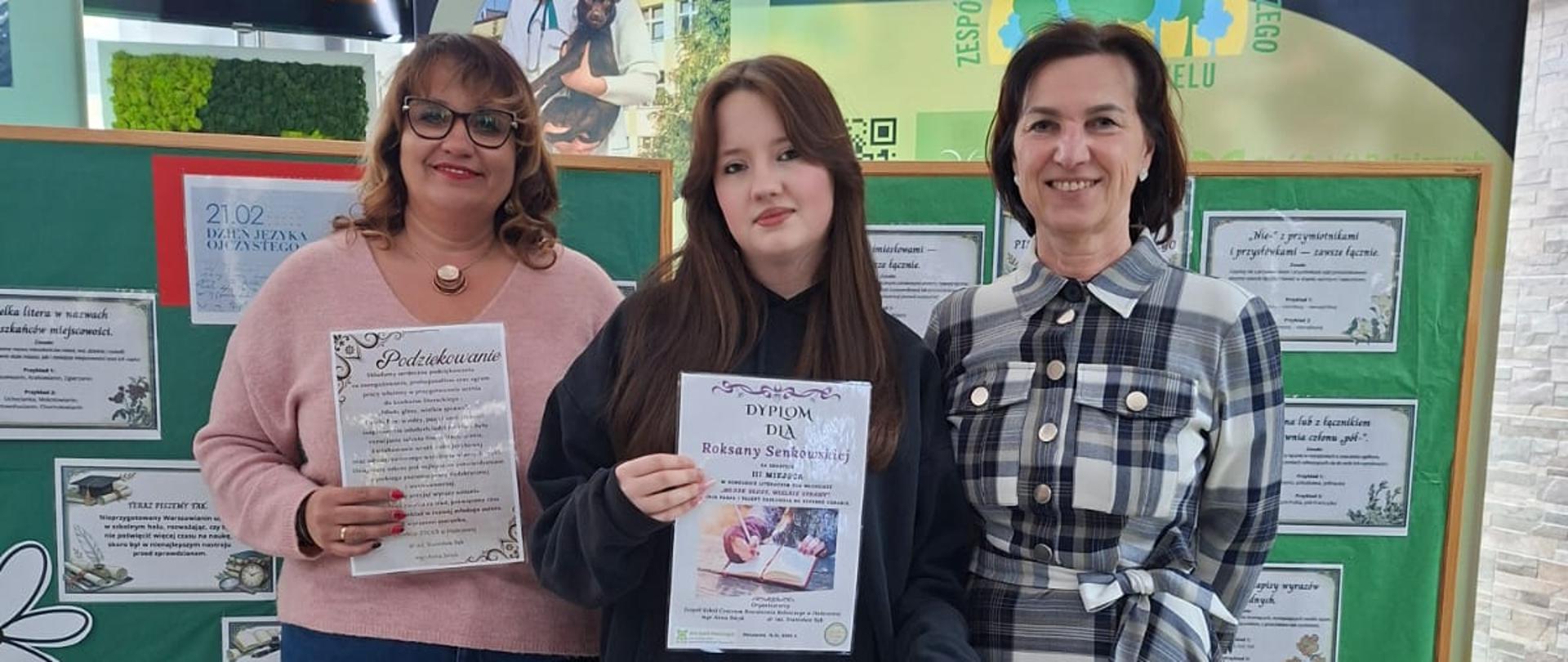 Trzy kobiety stoją na tle zielonej tablicy informacyjnej w szkolnym korytarzu. Pośrodku młoda dziewczyna, Roksana Senkowska, trzyma dyplom oraz białą torebkę prezentową. Po jej lewej stronie kobieta w różowym swetrze trzyma list z podziękowaniem, po prawej kobieta w długiej sukni w kratę. W tle widoczna tablica z logo Zespołu Szkół Centrum Kształcenia Rolniczego oraz gablota z pucharami.