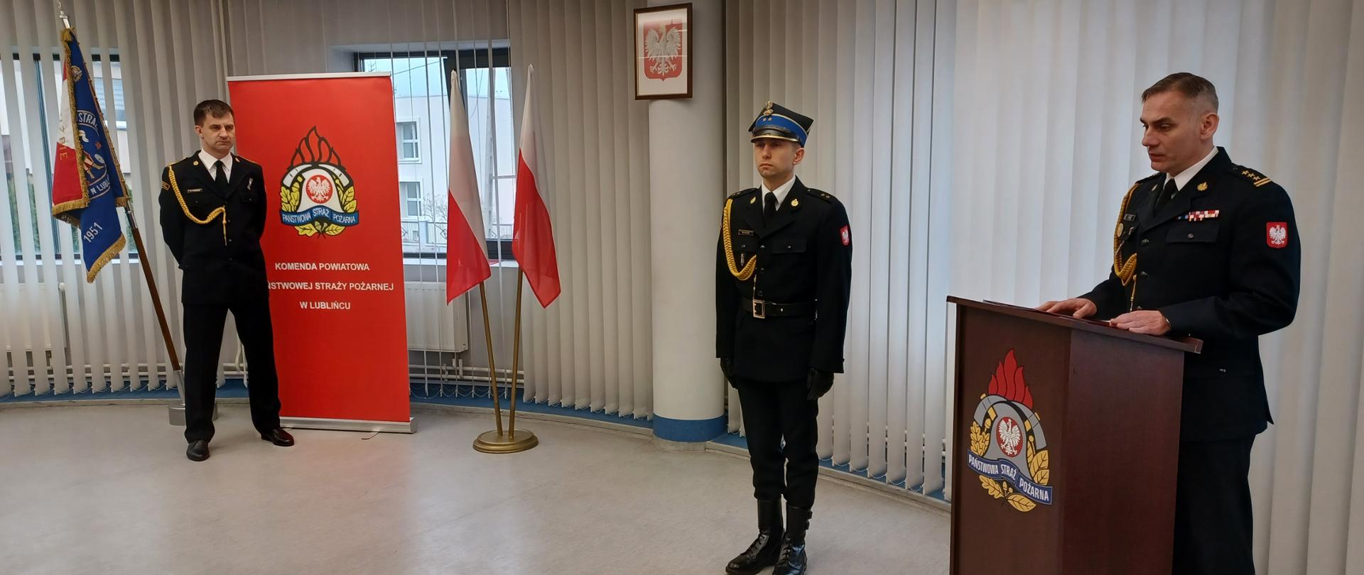 Zdjęcie zrobione w pomieszczeniu strażnicy na tle godła Rzeczpospolitej Polskiej, Sztandaru Komendy Powiatowej Państwowej Straży Pożarnej w Lublińcu, dwóch Flag Rzeczpospolitej Polskiej oraz baneru Komendy Powiatowej PSP w Lublińcu. Na pierwszym planie, po lewej stronie funkcjonariusz odchodzący na zaopatrzenie emerytalne, po prawej stronie Komendant Powiatowy Państwowej Straży Pożarnej w Lublińcu st. bryg. Janusz Bula stojący za mównicą oraz pomiędzy nim a flagami Rzeczpospolitej Polskiej dowódca uroczystości. Strażacy ubrani w umundurowanie galowe.