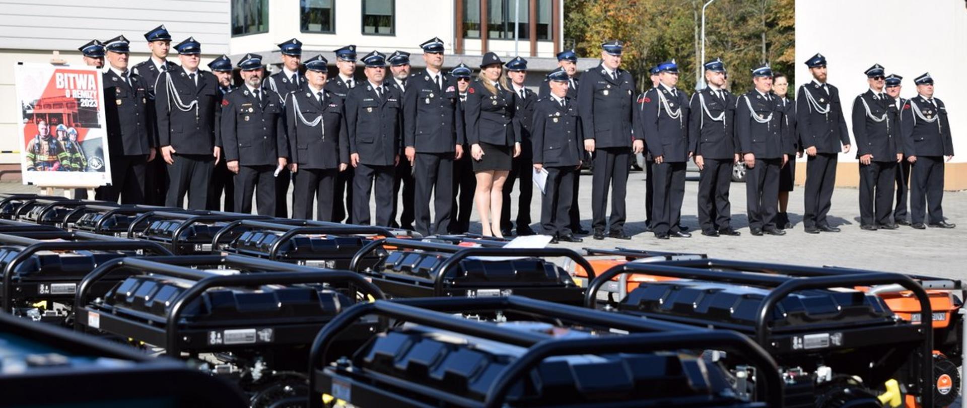 Zdjęcie zrobione na zewnątrz, placu Komendy Miejskiej Państwowej Straży Pożarnej w Rzeszowie, podczas uroczystego przekazania agregatów prądotwórczych ochotniczym strażom pożarnym. Druhny i druhowie Ochotnicy stoją w dwuszeregu przed nimi ustawione są agregaty prądotwórcze.
