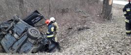 Samochód po dachowaniu leży poza drogą obok niego strażacy prowadzący działania. W tle zarośla.