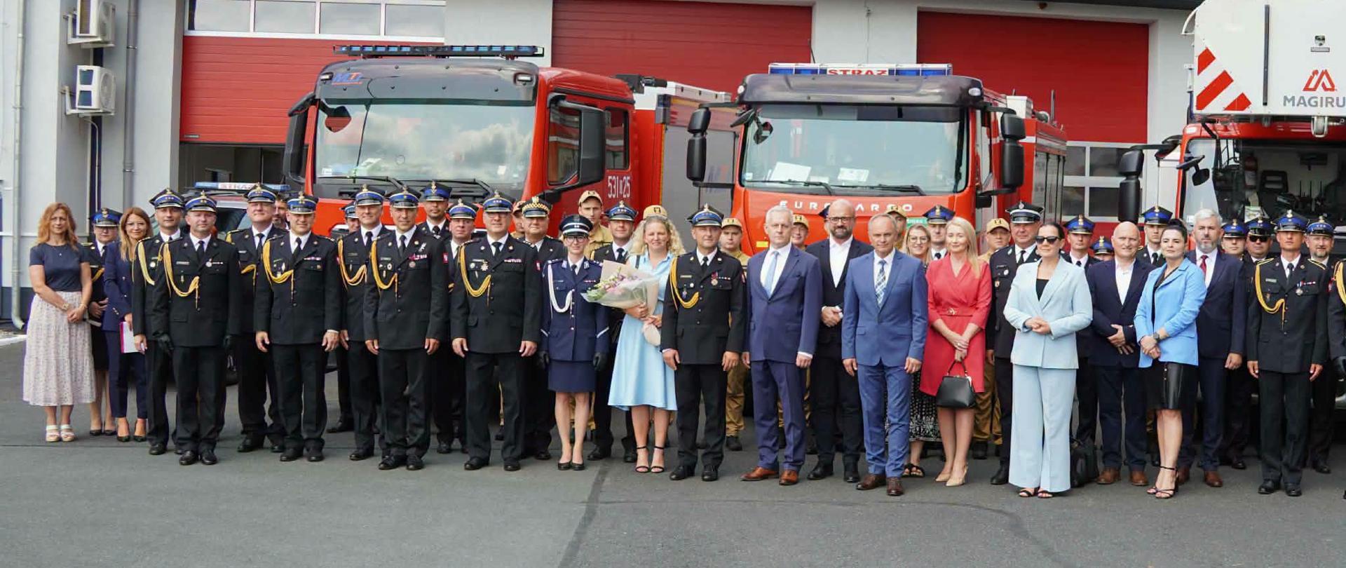 Grupowe zdjęcie pamiątkowe wykonane przed budynkiem Państwowej Straży Pożarnej. W tle widoczny jest nowoczesny budynek strażnicy z dużym napisem „PAŃSTWOWA STRAŻ POŻARNA” oraz godłem formacji nad trzema czerwonymi bramami garażowymi, w których zaparkowane są czerwone samochody ratowniczo-gaśnicze. Na pierwszym planie, na placu, stoi liczny rząd osób ustawionych do fotografii – większość z nich to strażacy w ciemnogranatowych mundurach wyjściowych i czapkach rogatywkach. W centralnej części grupy stoją zaproszeni goście oraz cywile w eleganckich strojach (garniturach i sukienkach), w tym kobieta w jasnoniebieskiej sukience trzymająca bukiet kwiatów.