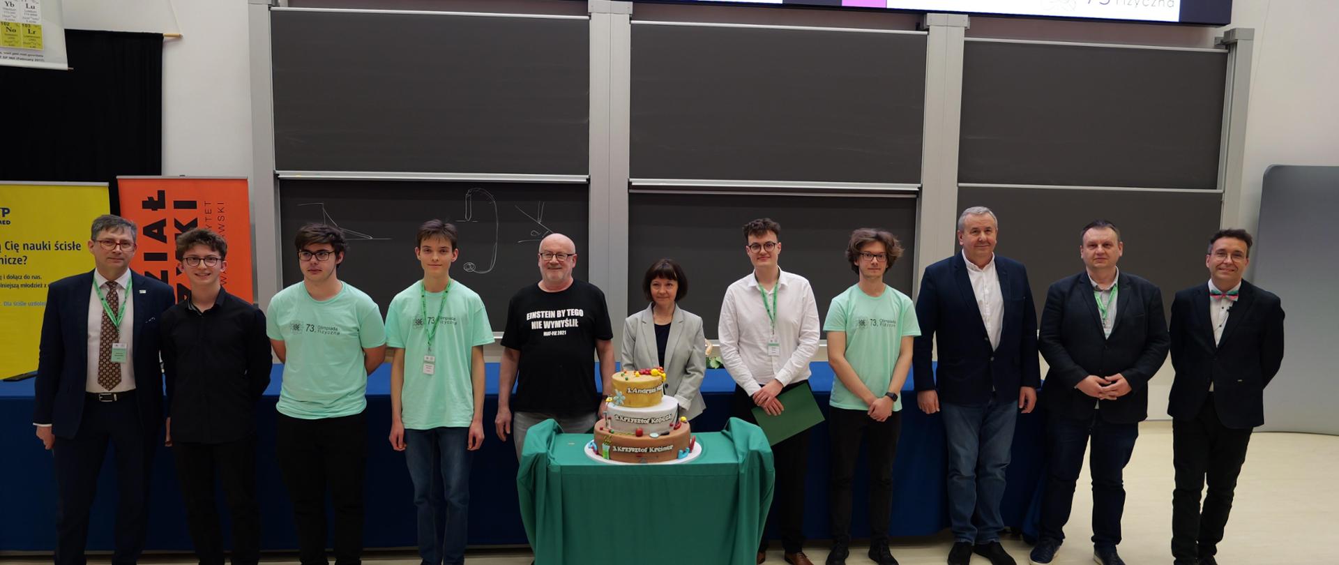 Zdjęcie zbiorowe laureatów i opiekunów 73 Olimpiady Fizycznej. Przed nimi na stoliku ustawiony jest trzy piętrowy tort z nazwiskami 3 laureatów