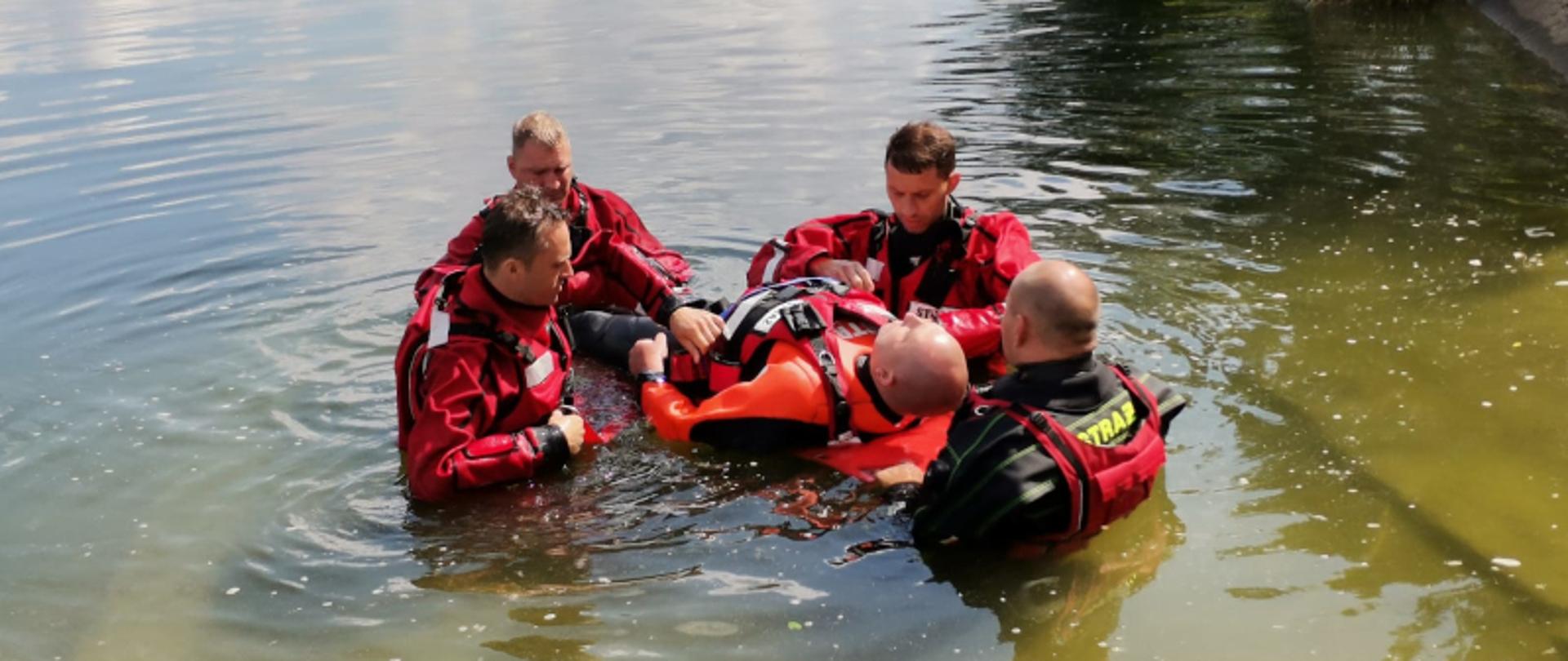 Czterech strażaków ubranych w czerwone skafandry do nurkowania podtrzymują na desce ratowniczej pozoranta