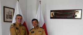 Strażacy w mundurze musztardowym uściskają sobie dłoń jeden z nich trzyma czerwoną teczkę za nimi stoją dwie flagi Polski na ścianach wiszą obraz oraz szabla. 