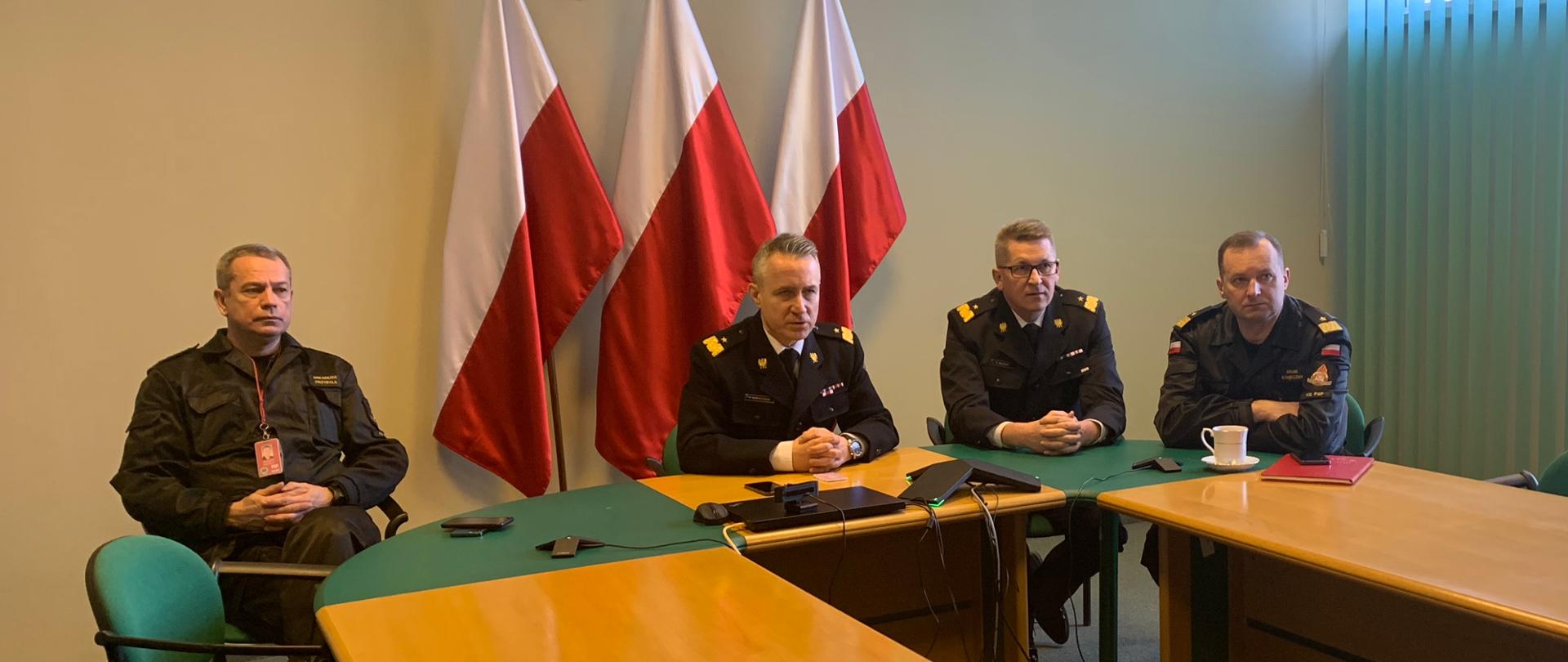 Kierownictwo Komendy Głównej Państwowej Straży Pożarnej podczas wideokonferencji. Za plecami uczestników flagi państwowe oraz godło państwowe