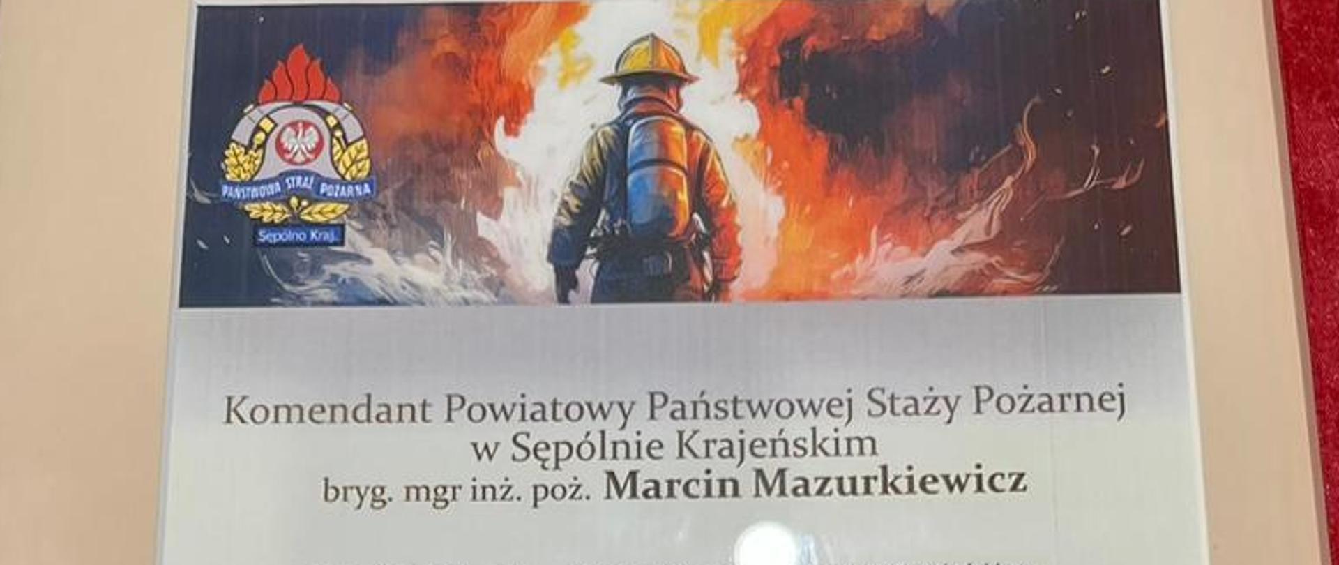 Życzenia z okazji Dnia Strażaka od włodarzy powiatu sępoleńskiego u góry rysunek przedstawiający strażaka na tle płomieni, całość oprawiona w ramkę i owinięte czerwoną tasiemką z kokardką.