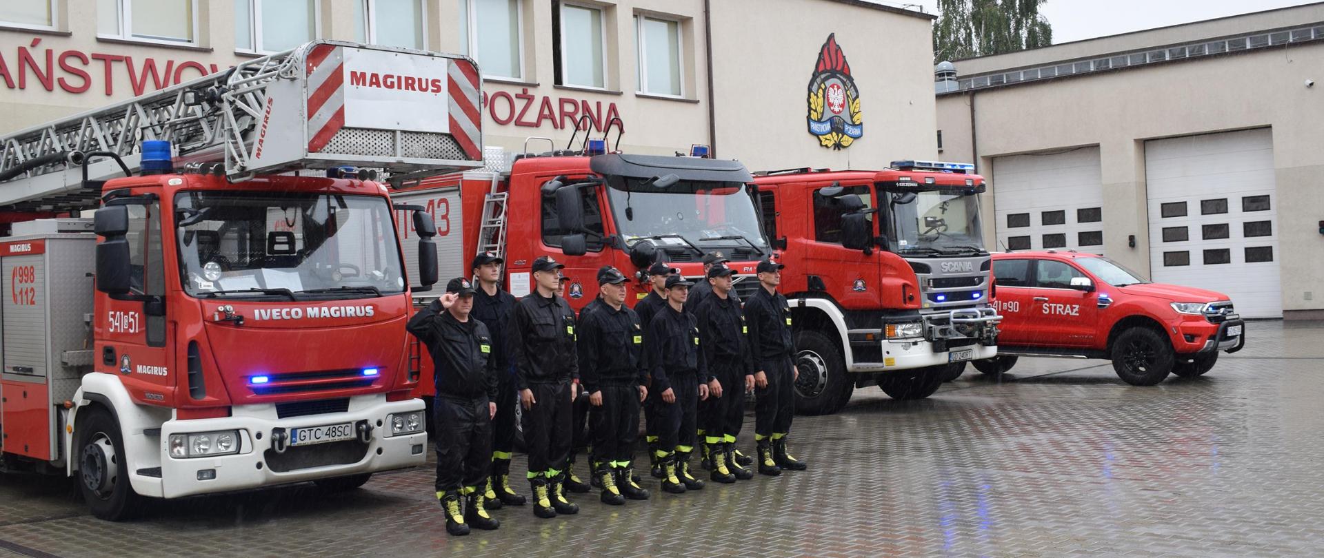 Plac JRG. 11 strażaków w umundurowaniu koszarowym stoi w dwuszeregu na baczność. Dowódca szyku oddaje honor do czapki. Za nimi samochody strażackie i budynek JRG.