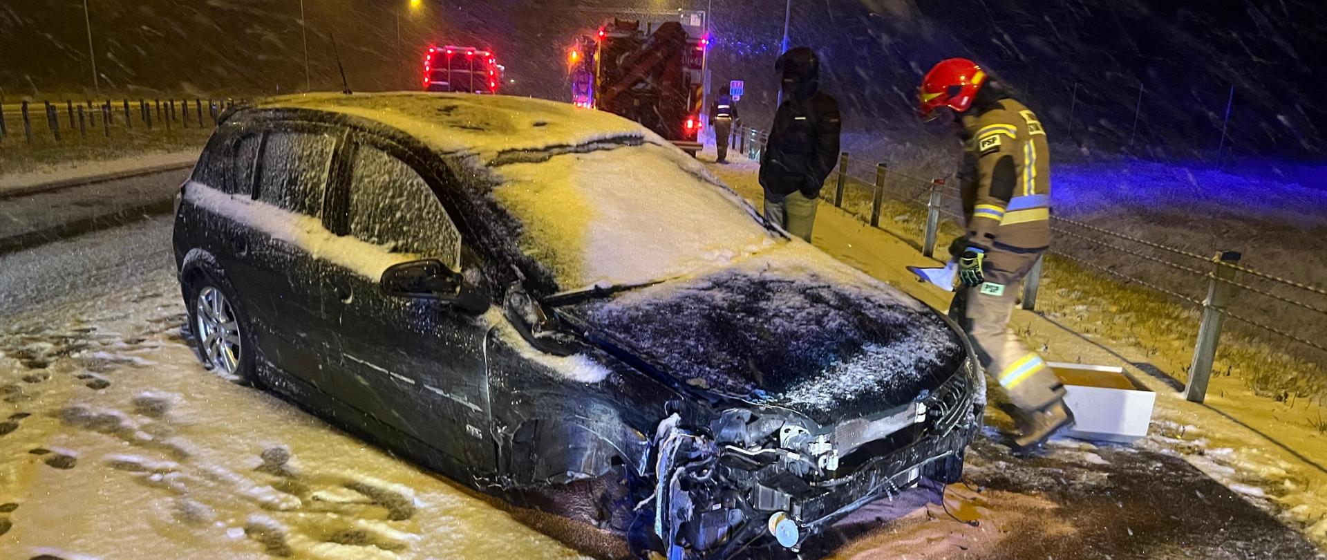 Na zdjęciu uszkodzony samochód osobowy na pasie awaryjnym trasy S7. Przy samochodzie strażak w umundurowaniu specjalnym, który rozsypuje sorbent celem neutralizacji płynów, które wyciekły z uszkodzonego pojazdu. W tle samochody pożarnicze. 