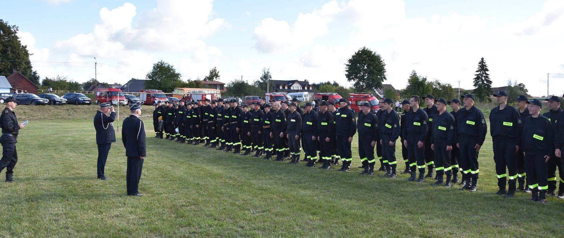Zdjęcie przedstawia strażaków ustawionych w szyku w tle pojazdy pożarnicze i cywilne