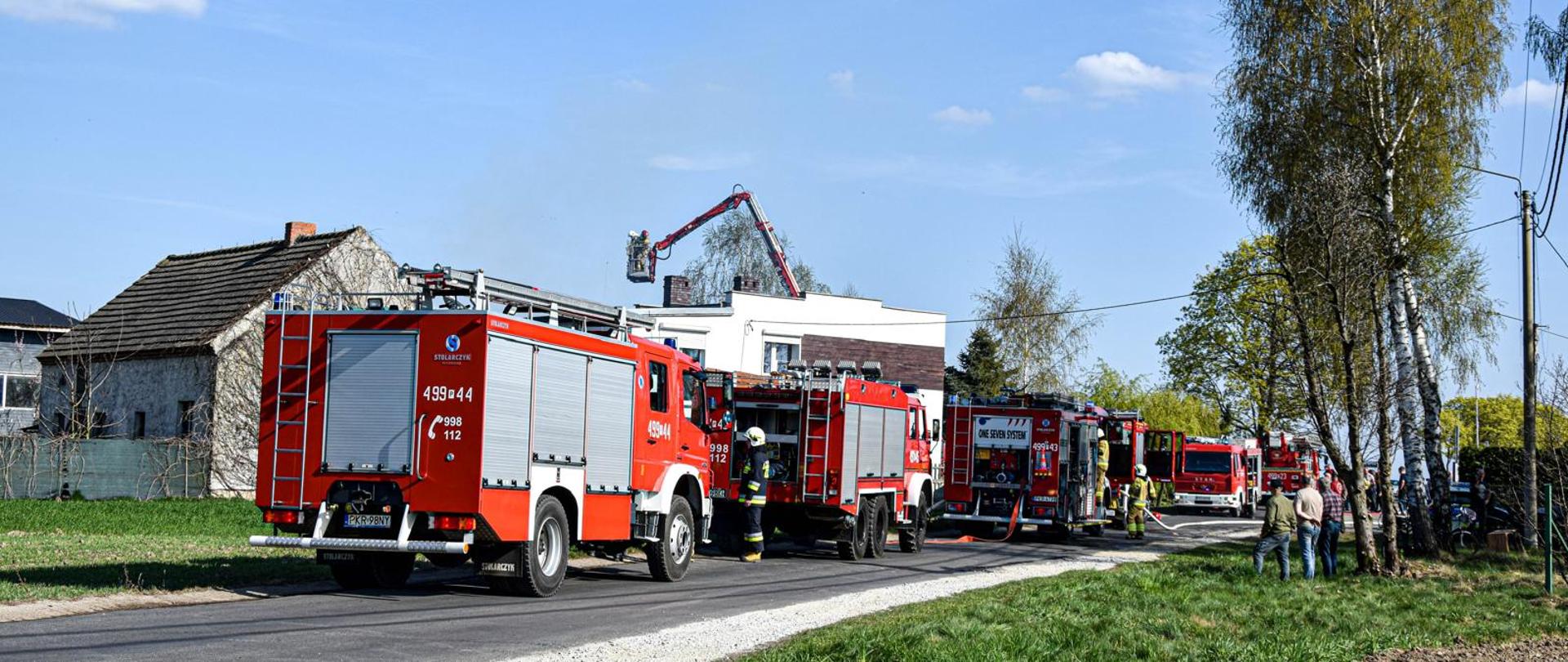 Na pierwszym planie widoczna kolumna pojazdów pożarniczych zaangażowana w działania gaśnicze związane z pożarem budynku mieszkalnego. 4 samochody stoją jeden za drugim na drodze. W oddali widoczny kosz podnośnika hydraulicznego. 