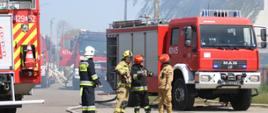 Zdjęcie przedstawia strażaków, samochody pożarnicze oraz rozwinięte węże pożarnicze na ulicy Mickiewicza przed bramami wjazdowymi na teren zakładów. 