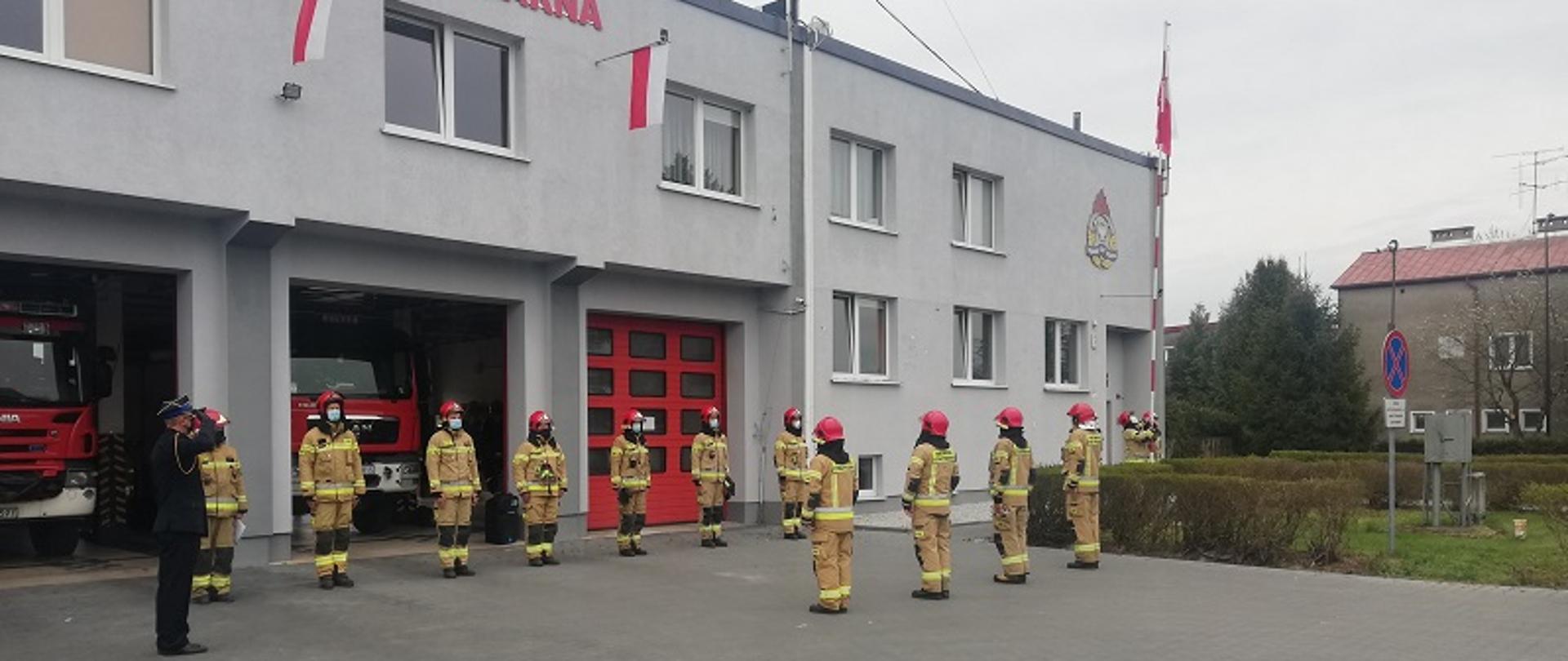 Zdjęcie przedstawia uroczystą zmianę służby podczas obchodów Święta Flagi. Dwa szeregi strażaków w umundurowaniu specjalnym koloru piaskowego i czerwonymi hełmami stoją w odstępach naprzeciwko siebie. Pośrodku szeregów salutujący st. kpt. Mariusz Wenta z-ca dowódcy JRG w umundurowaniu wyjściowym. Strażacy na twarzach mają założone maseczki ochronne. W tle widoczny budynek strażnicy. Po prawej strażak wciąga flagę państwową na maszt