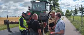 Ciągnik rolniczy stoi na drodze obok dwie osoby cywilne, policjant i strażak.