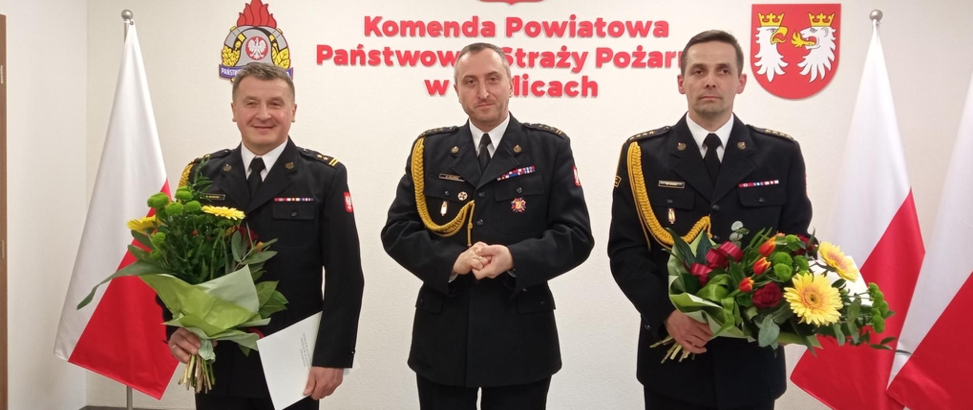 Na zdjęciu w mundurach galowych koloru granatowego na sali konferencyjnej Komendy Powiatowej Państwowej Straży Pożarnej w Gorlicach podczas uroczystej zbiórki stoi trzech strażaków. Po lewej odchodzący komendant powiatowy Państwowej Straży Pożarnej w Gorlicach w środku małopolski komendant wojewódzki Państwowej Straży Pożarnej a po prawej nowy komendant powiatowy Państwowej Straży Pożarnej w Gorlicach. Za ich plecami na ścianie napis koloru czerwonego Komenda Powiatowa Państwowej Straży Pożarnej w Gorlicach godło logo PSP i Godło powiatu gorlickiego i flagi biało-czerwone.