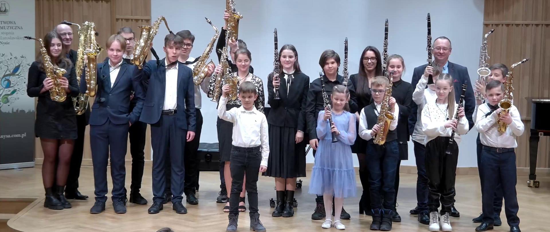 Zdjęcie. Na estradzie Sali Koncertowej PSM I i II stopnia w Nysie stoją w grupie wszyscy wykonawcy - uczniowie i nauczyciele. Saksofoniści i klarneciści prezentują swoje instrumenty wznosząc je do góry. W dolnej części zdjęcia widać słuchaczy, którzy telefonami komórkowymi robią zdjęcia zebranym na estradzie.
