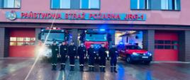 Na zdjęciu widzimy siedmiu strażaków stojących w szeregu. Ubrani są w mundury koszarowe koloru czarnego. Na głowach czarne czapki z orłem na środku. strażak z lewej strony salutuje. Strażacy znajdują się przed budynkiem jednostki, za nimi widzimy samochody strażackie koloru czerwonego. Samochody mają załączone światła ostrzegawcze koloru niebieskiego. 