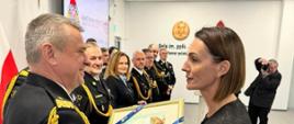 Strażak w mundurze wyjściowym ze sznurem otrzymuje o kobiety obraz z niebieskimi wstążkami obok stoją strażacy rodziny oraz zaproszeni goście.