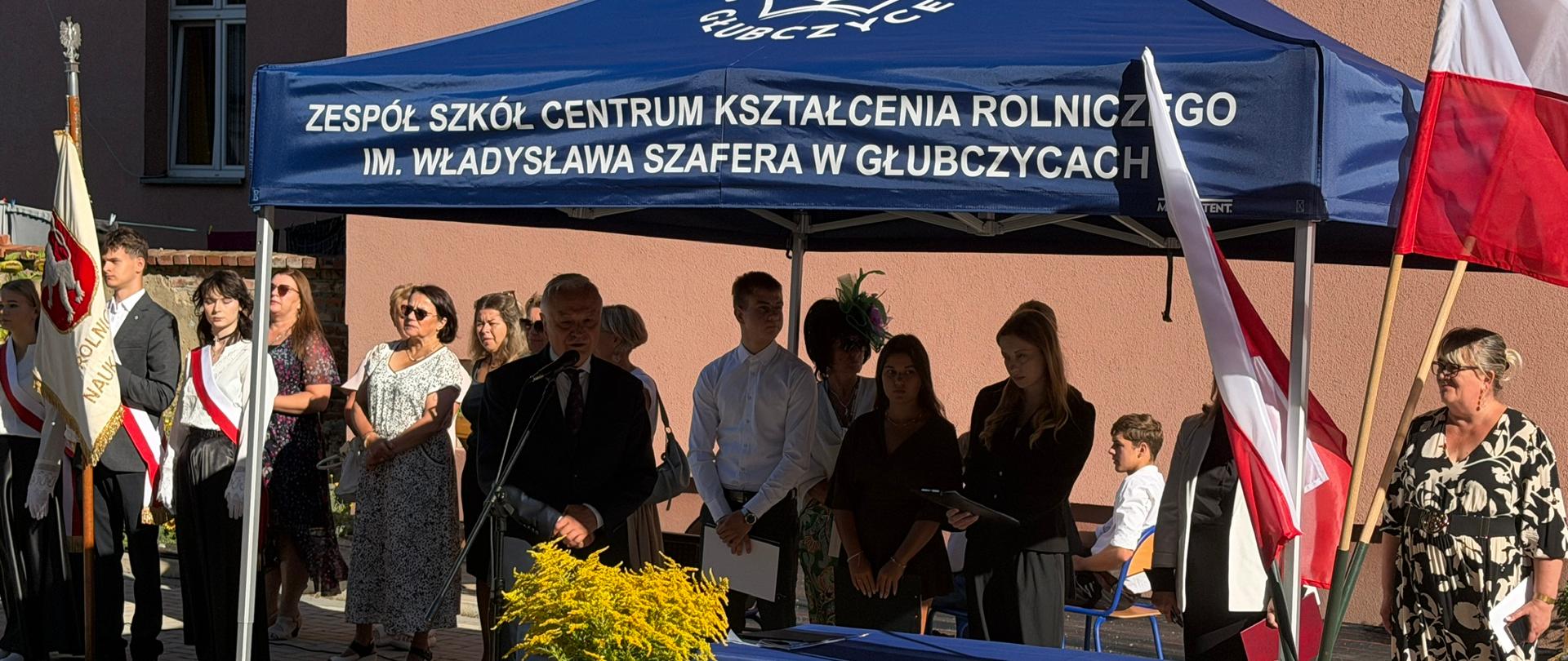 Dyrektor szkoły podczas uroczystego rozpoczęcia roku szkolnego w obecności uczniów, nauczycieli i pocztu sztandarowego.