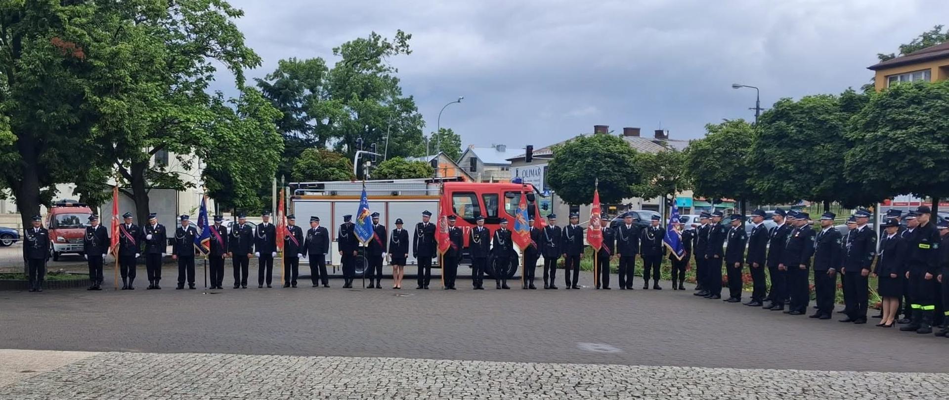 Szerokie ujęcie pokazuje strażaków ustawionych w długim szeregu podczas uroczystego apelu na Placu Stary Rynek. Funkcjonariusze stoją na brukowanej nawierzchni w galowych mundurach, zachowując równy szyk. W tle widoczne są drzewa, pojazdy strażackie oraz budynki miasta, a nad całością rozciąga się zachmurzone niebo.