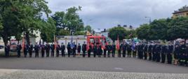 Szerokie ujęcie pokazuje strażaków ustawionych w długim szeregu podczas uroczystego apelu na Placu Stary Rynek. Funkcjonariusze stoją na brukowanej nawierzchni w galowych mundurach, zachowując równy szyk. W tle widoczne są drzewa, pojazdy strażackie oraz budynki miasta, a nad całością rozciąga się zachmurzone niebo.
