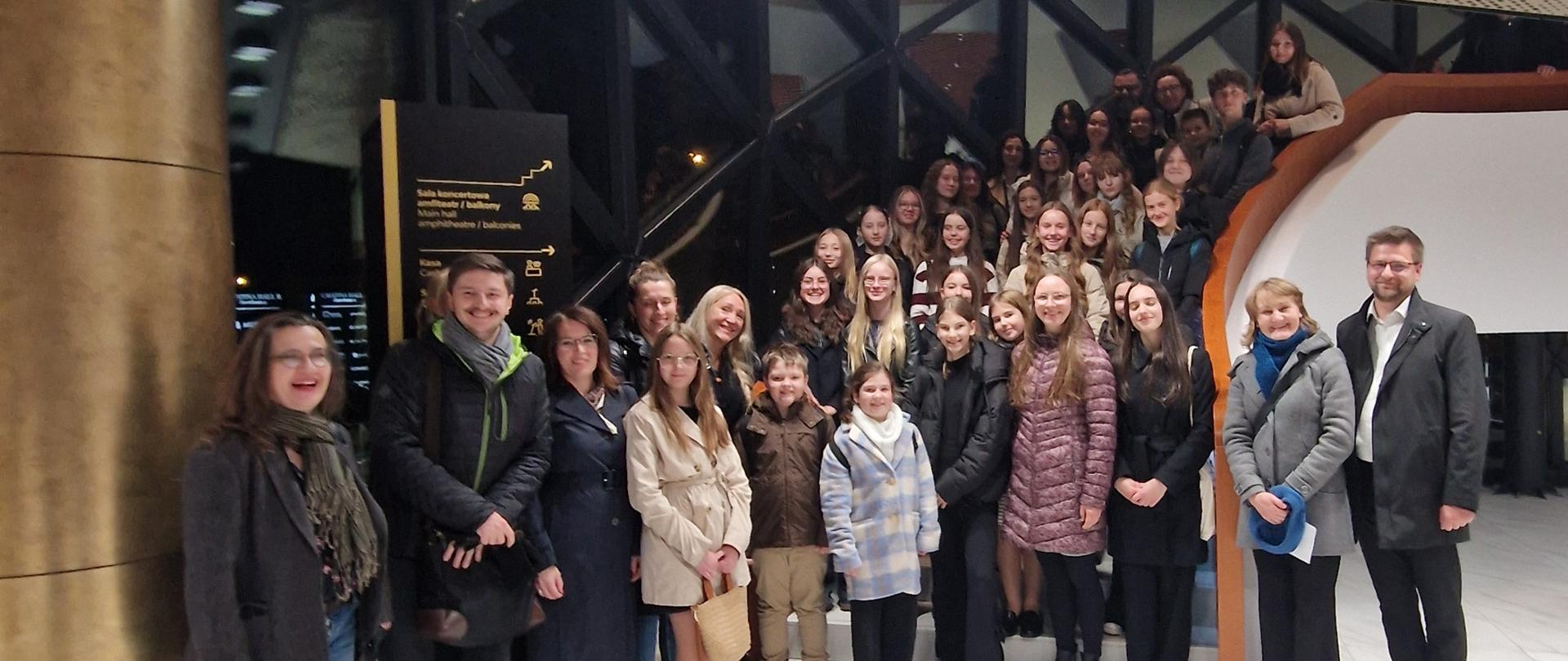 Na zdjęciu widać dużą grupę ludzi – około czterdziestu osób – stojących razem we wnętrzu nowoczesnego budynku. Pomieszczenie jest jasne, z marmurową podłogą i złotymi, błyszczącymi elementami wystroju. W tle widać czarną, metalową konstrukcję i przeszklone ściany. Część osób stoi na podłodze, a część – na schodach z prawej strony, dzięki czemu wszyscy dobrze się mieszczą w kadrze.
W grupie są głównie młode kobiety i dziewczęta, ale też kilku chłopców i dorosłych – nauczycieli. Większość jest ciepło ubrana w płaszcze, kurtki i szaliki, co sugeruje, że zdjęcie zrobiono jesienią lub zimą. Wszyscy patrzą w stronę aparatu i uśmiechają się – atmosfera jest serdeczna i zorganizowana, jak podczas wspólnego wyjścia grupy szkolnej lub chóru.