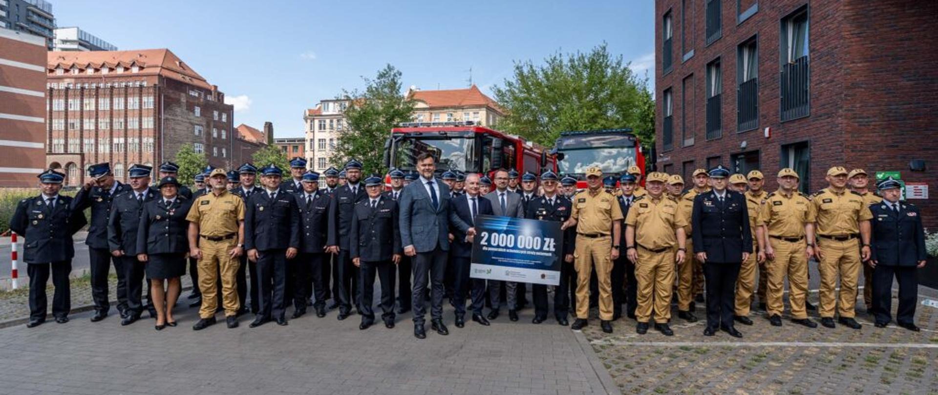 Funkcjonariusz Państwowej Straży Pożarnej zaproszeni goście oraz strażacy ochotniczych straży pożarnych stoją obok siebie na tle dwóch samochodów gaśniczych w środku grupy jest napis dwa miliony złotych.