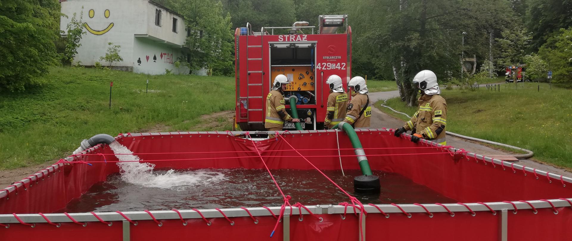 Zdjęcie przedstawia samochód pożarniczy stojący przy rozkładanym zbiorniku wodnym o pojemności 13 metrów sześciennych. Przy zbiorniku soją strażacy którzy obsługują przesyłanie wody na dalsze odległości.