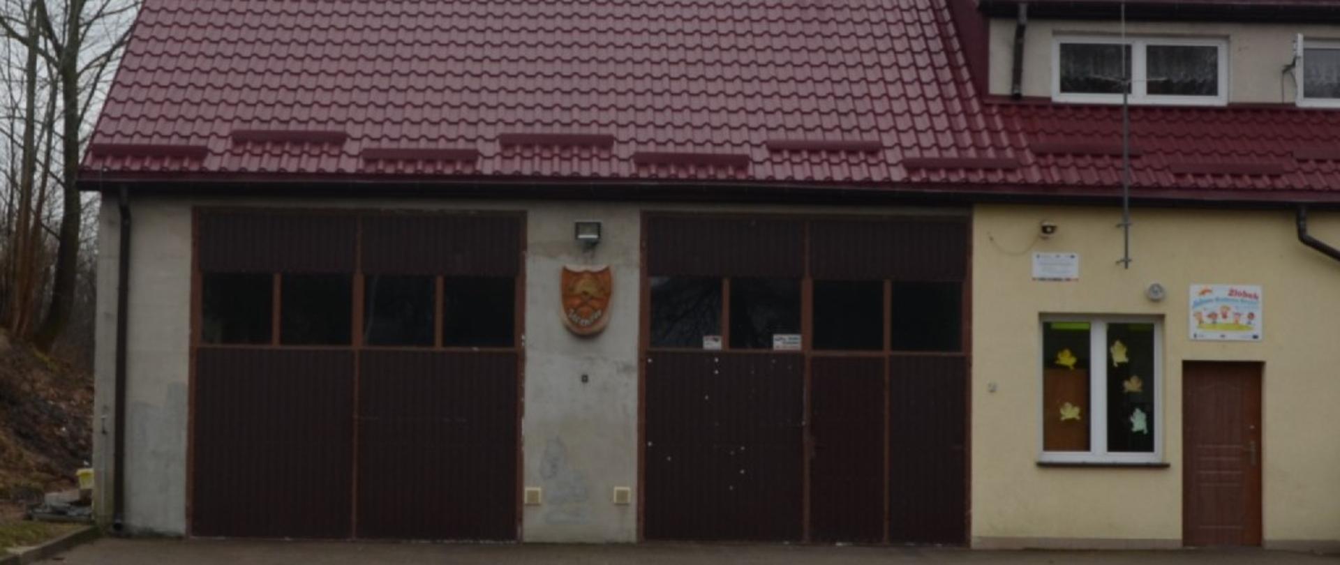 Widoczny jest budynek z dachem koloru czerwonego. Dwie bramy koloru brązowego. Po prawej stronie drzwi wejściowe do zaplecza socjalnego remizy. Przed budynkiem podjazd z kostki brukowej .