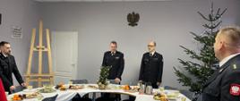 Przy zastawionym stole stoją Komendanci Powiatowi PSP w Bytowie w galowych mundurach. Z prawej strony stoi żywa choinka z niebieskimi lampkami
