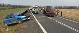 Na zdjęciu widoczne są uszkodzone po wypadku samochody : samochód osobowy i dostawczy. Samochód osobowy znajduje się na poboczu drogi. W tle widoczna jest karetka pogotowia oraz samochody straży pożarnej. 