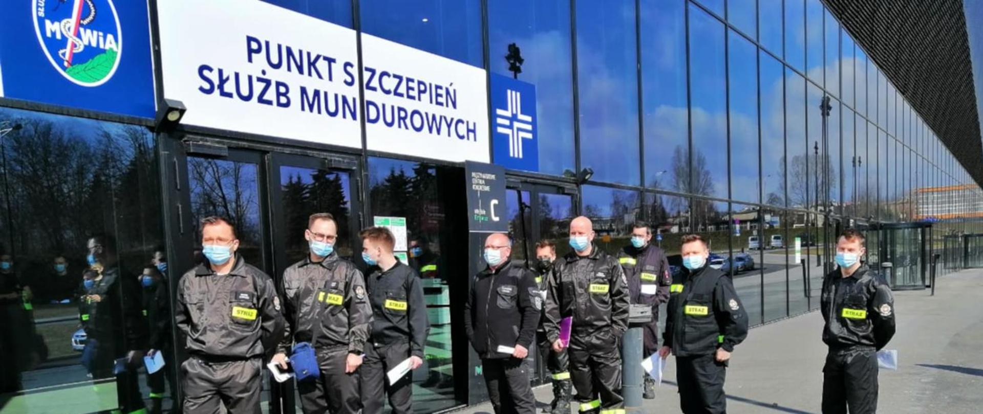 Zdjęcie przedstawia 9 strażaków ubranych w ubraniu koszarowym stojących przed drzwiami budynku MCK w Katowicach w kolejce do szczepienia. Nad drzwiami widnieje napis Punkt Szczepień Służb Mundurowych.