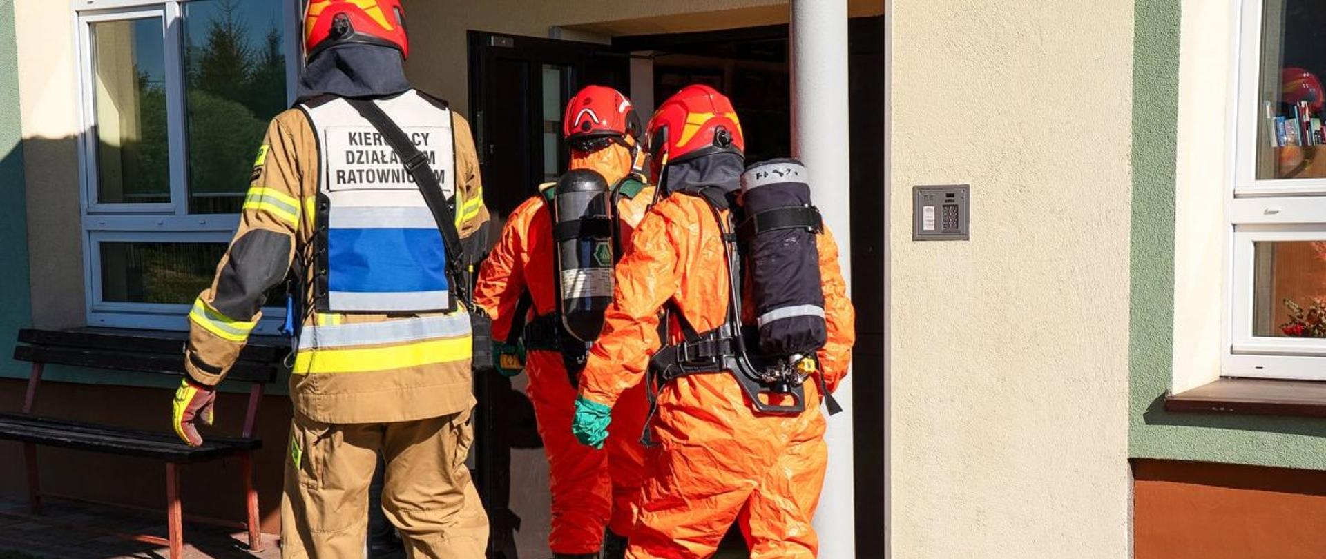 Zdjęcie przedstawia Kierującego Działaniem Ratowniczym oraz strażaków w ubraniach chemoodpornych którzy wchodzą do środka szkoły przez wejście główne. 