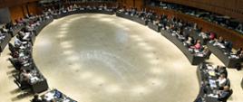 Widok z góry - wielka sala, okrągły stół przy którym siedzi wiele osób przed monitorami.