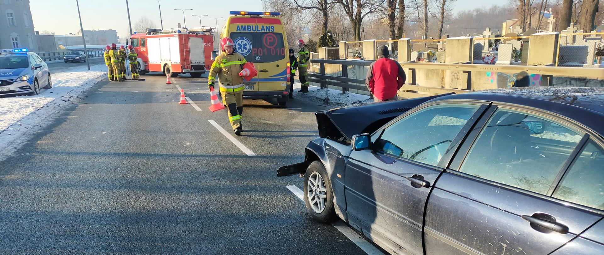 Na zdjęciu widać jak strażak na drodze dwupasmowej rozkłada pachołki służące do oznakowania terenu akcji. Po prawej stronie widać rozbity pojazd z uszkodzonym przodem. W tle stoi karetka pogotowia oraz samochód pożarniczy przy którym stoją strażacy