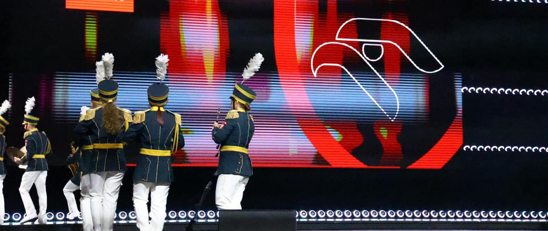 Na scenie kilkoro muzyków orkiestry w strojach okolicznościowych, z prawej stronie, na tle, logo firmy. Wokół światła estradowe.