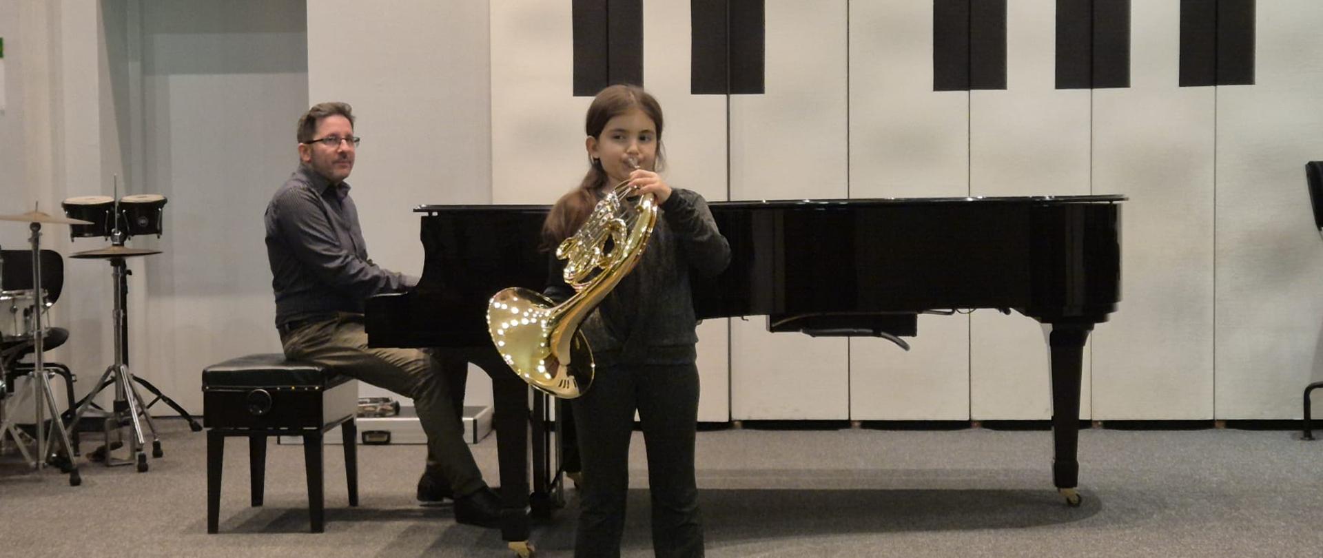 Dziewczynka gra na waltorni, mężczyzna akompaniuje jej na fortepianie.